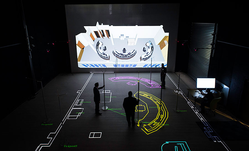 Large empty dark room, with brightly coloured lined shapes projected onto the floor. The shapes are outlines of office furniture, demonstrating the layout of an office space. Large empty dark room, with brightly coloured lined shapes projected onto the floor. The shapes are outlines of office furniture, demonstrating the layout of an office space.