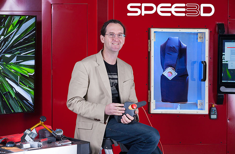 Person sitting from of large metal box machine the size of a small room. It has a window in one side showing a fabric covered robotic arm. Person sitting from of large metal box machine the size of a small room. It has a window in one side showing a fabric covered robotic arm.