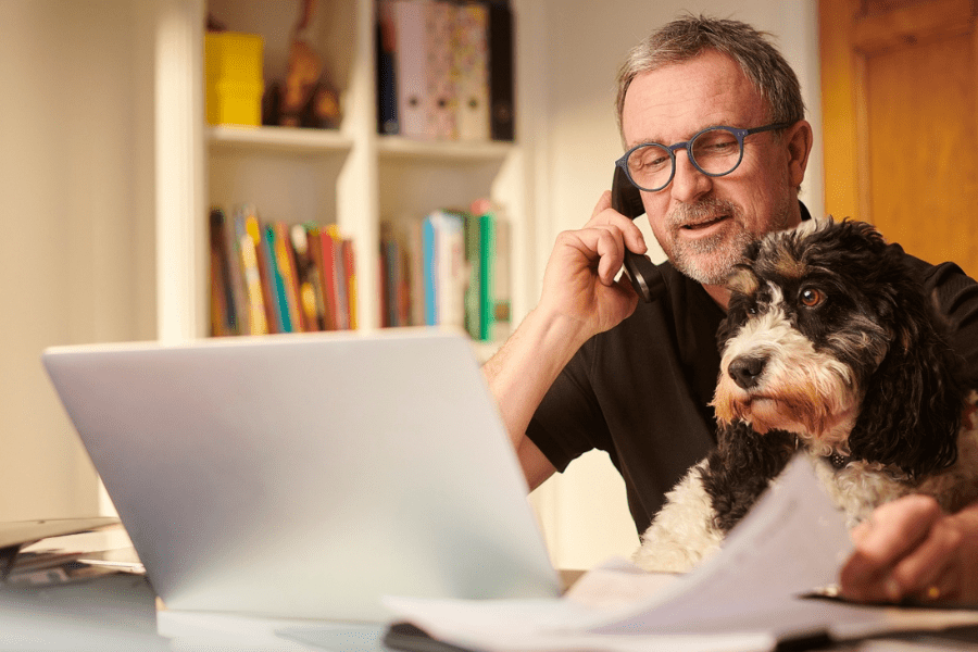 A man looks at his laptop while talking on the phone next to his dog