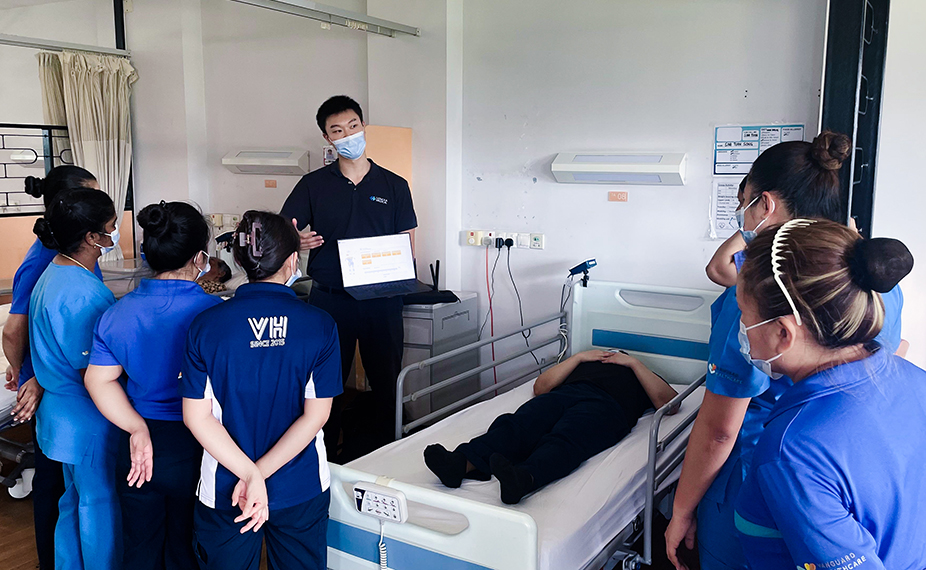 Image of a patient in a hospital bed with medical equipment; a person beside them is reading (LenexaCARE) data from a monitor.