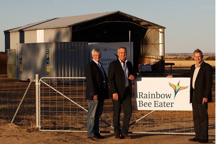 Three men standing at a farm gate with a Rainbow Bee Eater sign on it and a large shed in the background.
