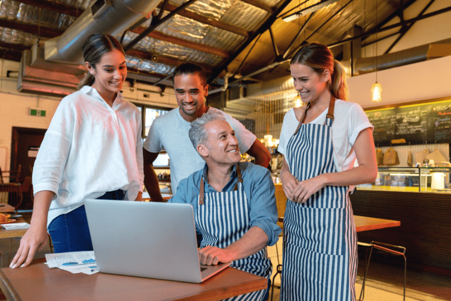 Four restaurant employees gather around a table at their restaurant and look at a laptop.