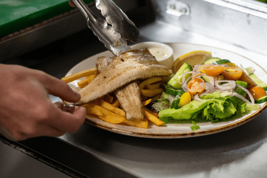 A close up of a plate of grilled fish, chips and salad being prepared.