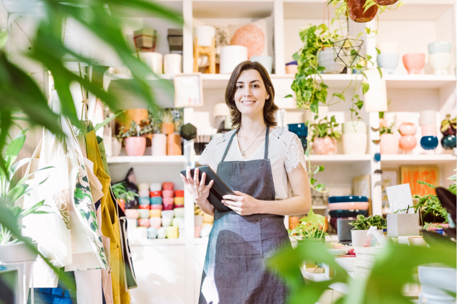 A smiling woman in an apron stands in a retail store filled with plants and homewares/